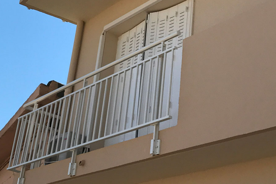Bâtiment beige avec balustrade de balcon blanche, volets et fond de ciel bleu.