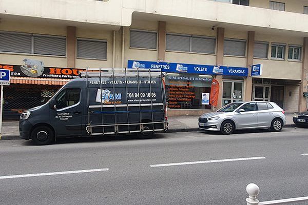 Une camionnette avec des supports pour le verre garée dans une rue de la ville devant un magasin avec un auvent bleu.