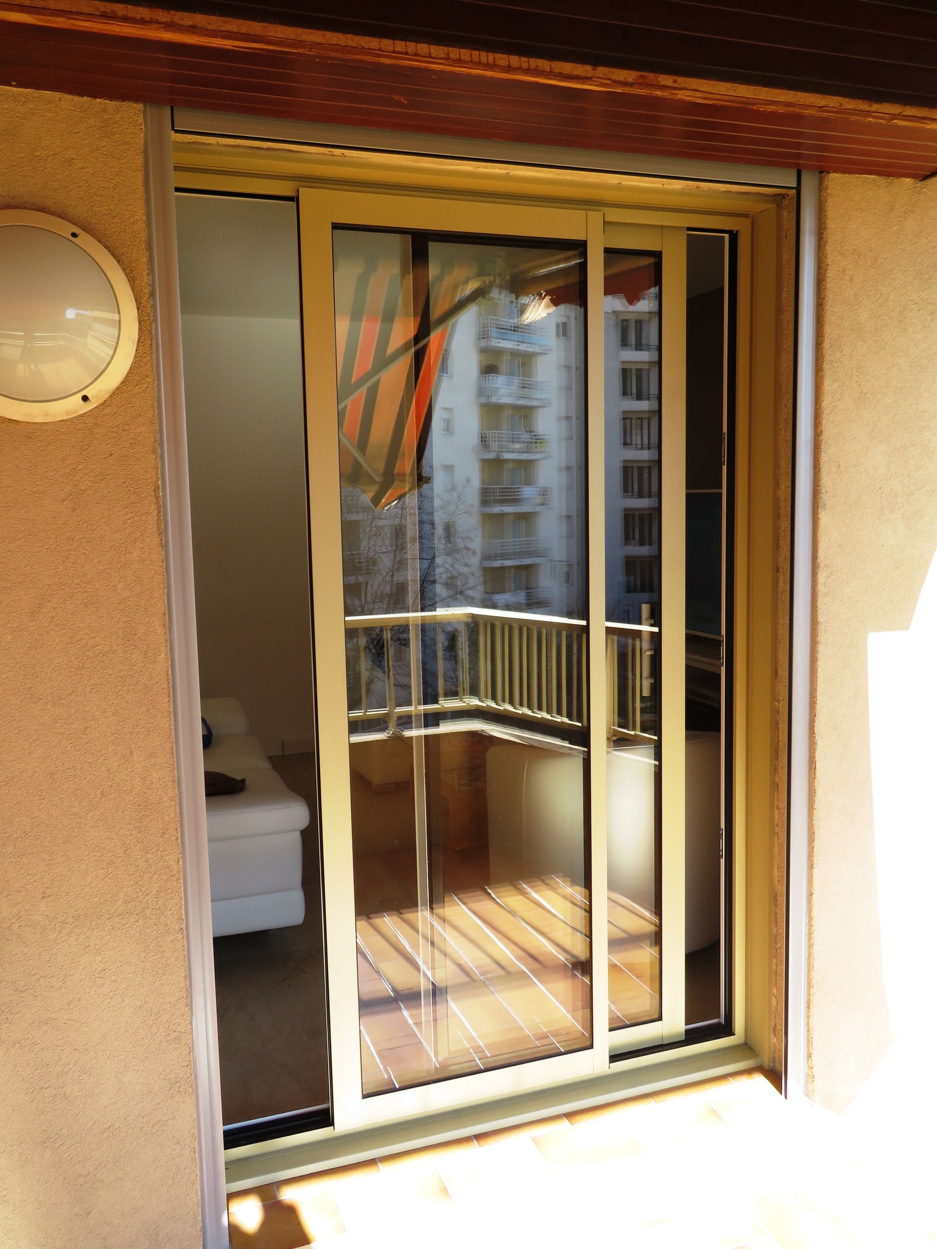 Porte-fenêtre coulissante en verre, cadre beige, ouvrant sur un balcon avec parquet et garde-corps. Bâtiment en arrière-plan.