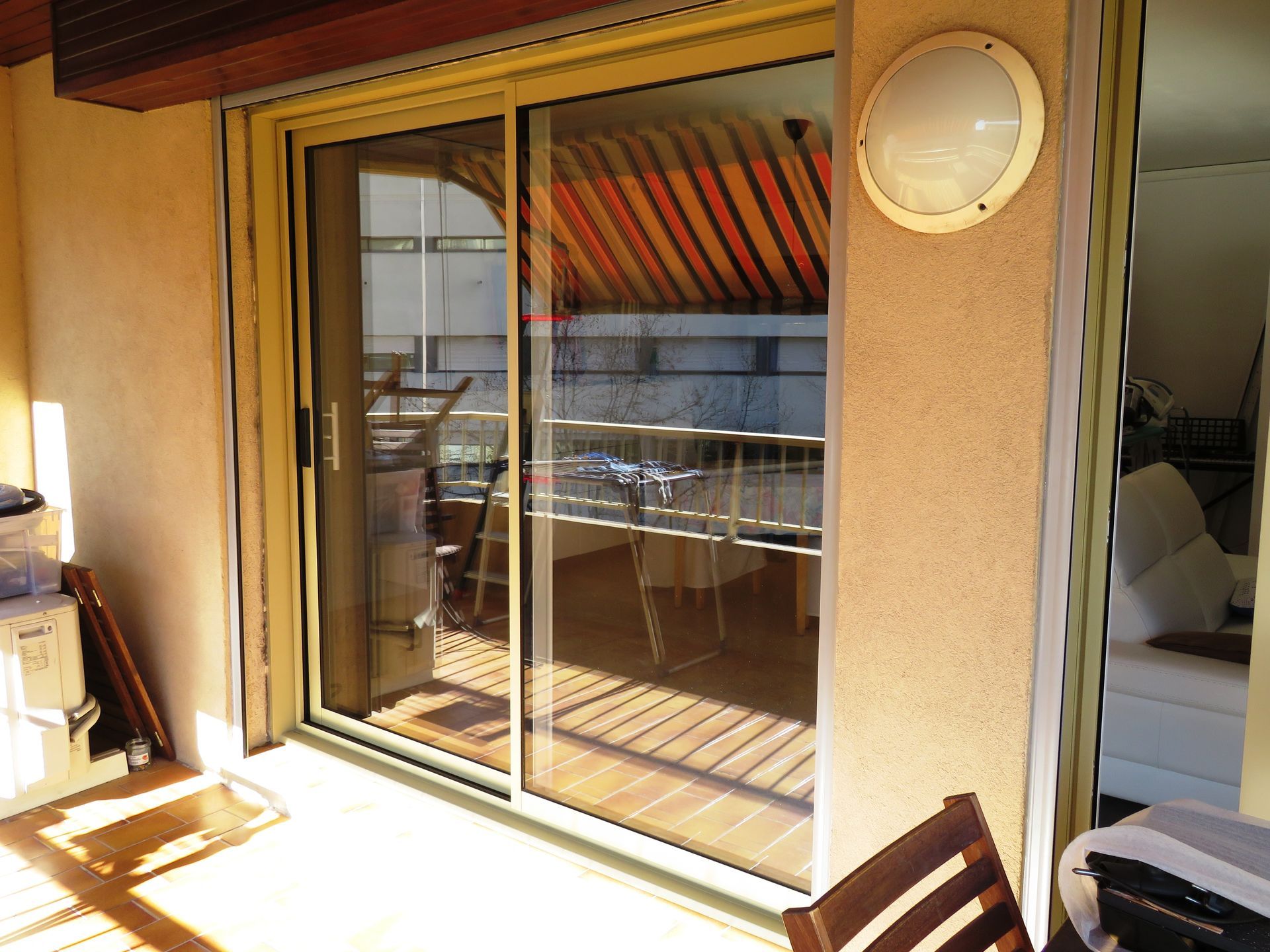 Porte coulissante en verre donnant sur un balcon avec balustrade en bois, murs beiges et luminaire circulaire.