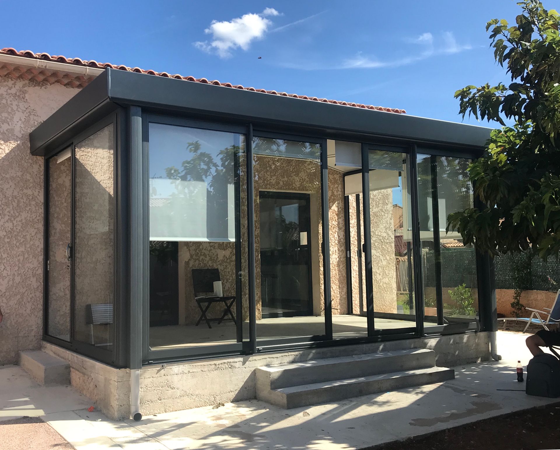 Véranda aux parois de verre avec un toit et une charpente gris foncé, construite sur une maison en stuc de couleur claire, avec des marches qui y mènent.