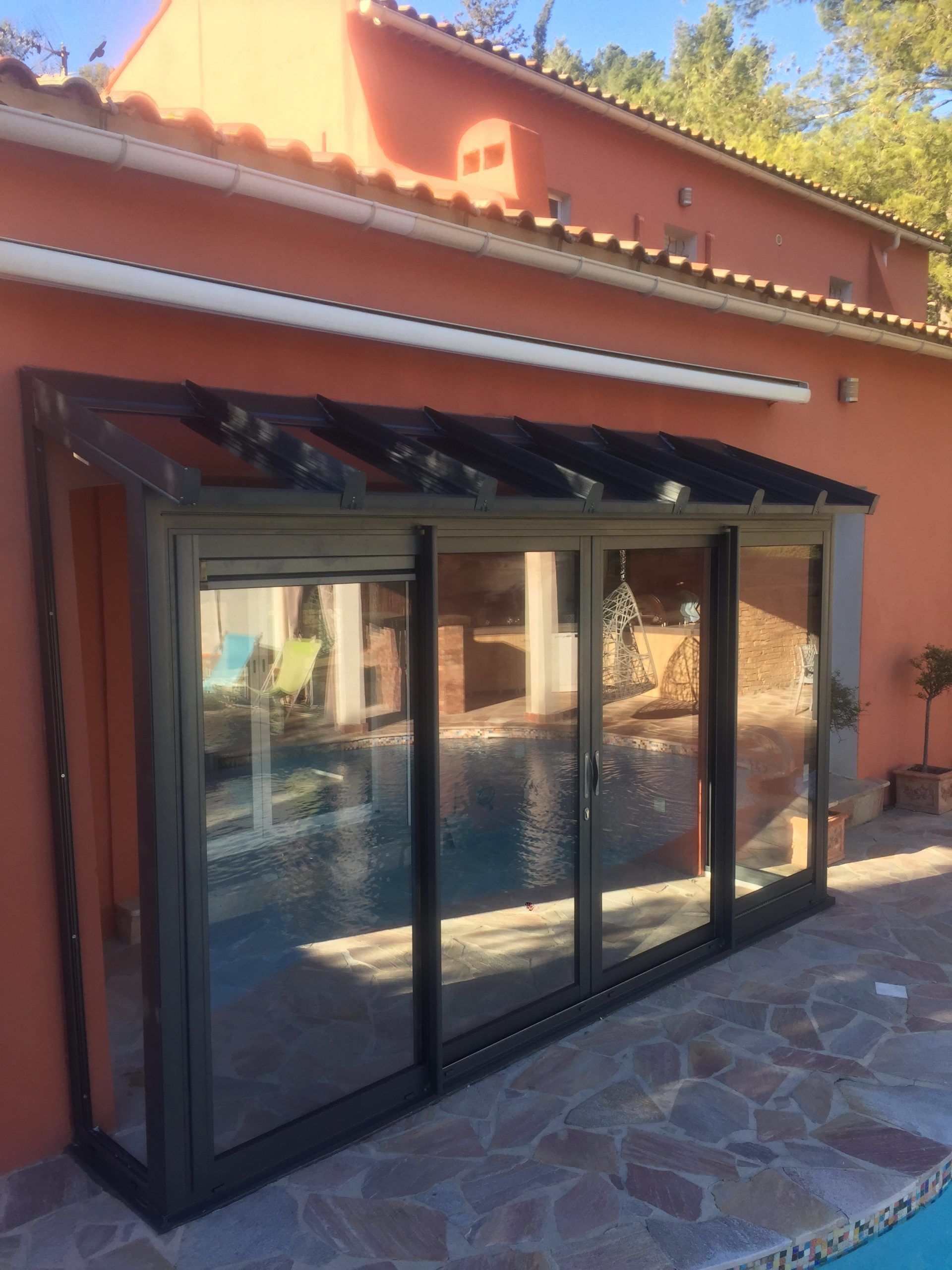 Abri de piscine avec cadre noir et panneaux en verre transparent, fixé à un bâtiment en stuc orange.