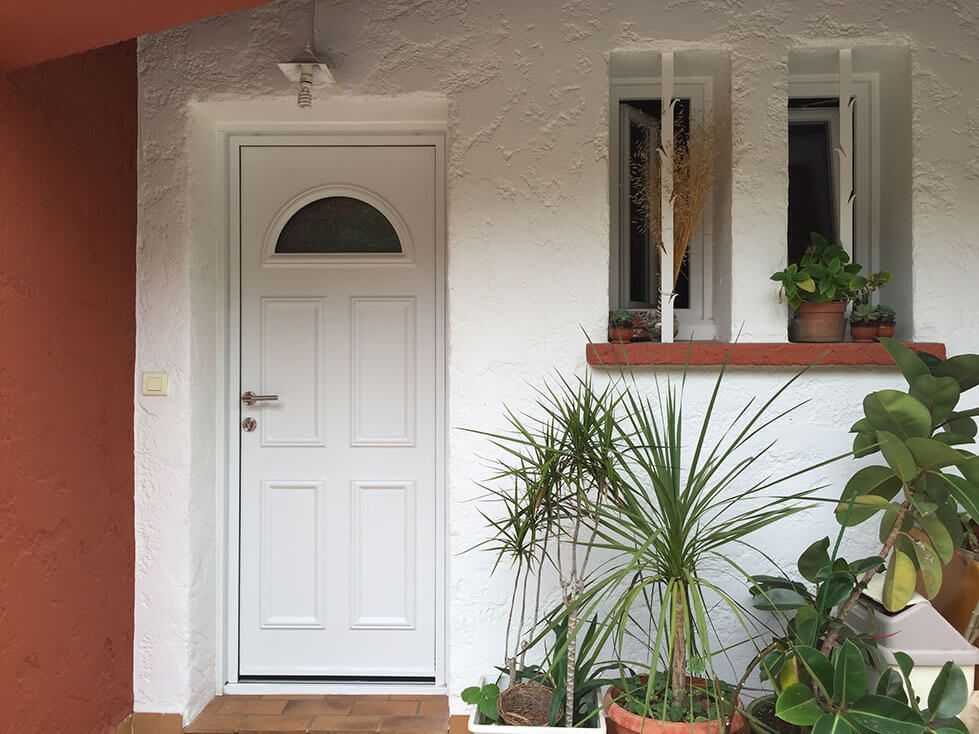 Porte blanche avec verre arqué au-dessus des panneaux, à côté d'une fenêtre et de plantes en pot.