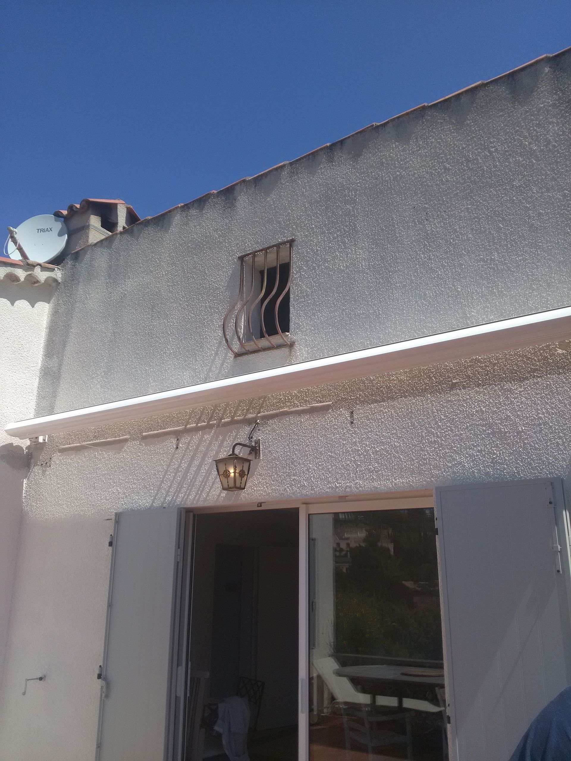 Vue extérieure d'un bâtiment en stuc avec une porte coulissante en verre et une petite fenêtre à barreaux sous un ciel bleu.