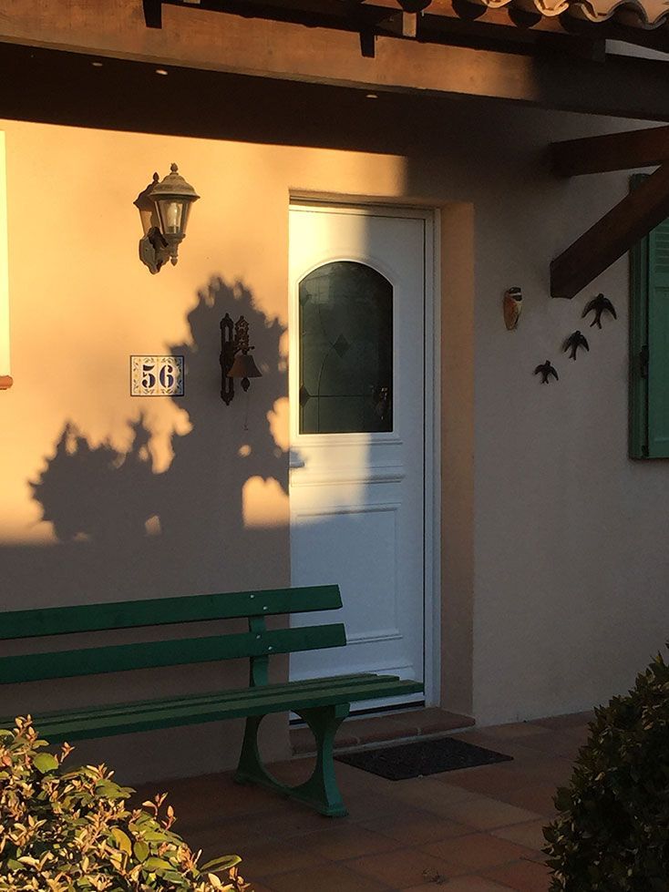 Extérieur d'un bâtiment avec une porte blanche, un banc vert et l'ombre d'un arbre sur le mur.