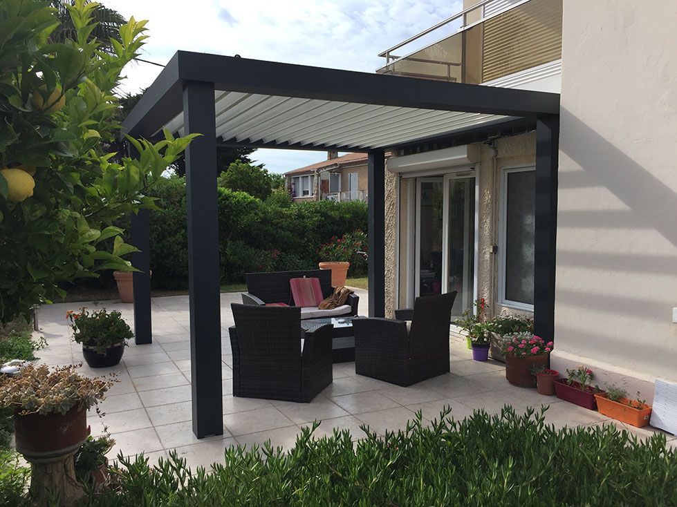 Terrasse avec pergola gris foncé surplombant un espace extérieur. Une végétation luxuriante entoure l'espace carrelé, à côté d'un bâtiment.