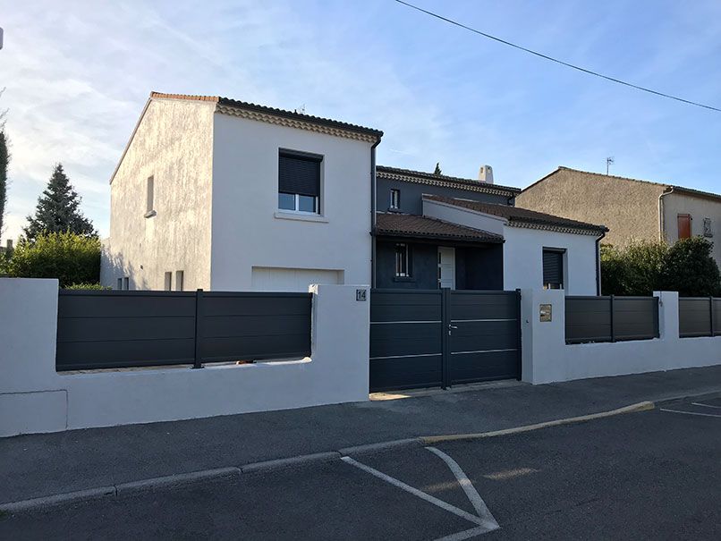Maison à deux étages avec clôture et portail gris, dans une rue résidentielle. Murs blancs, ciel bleu.