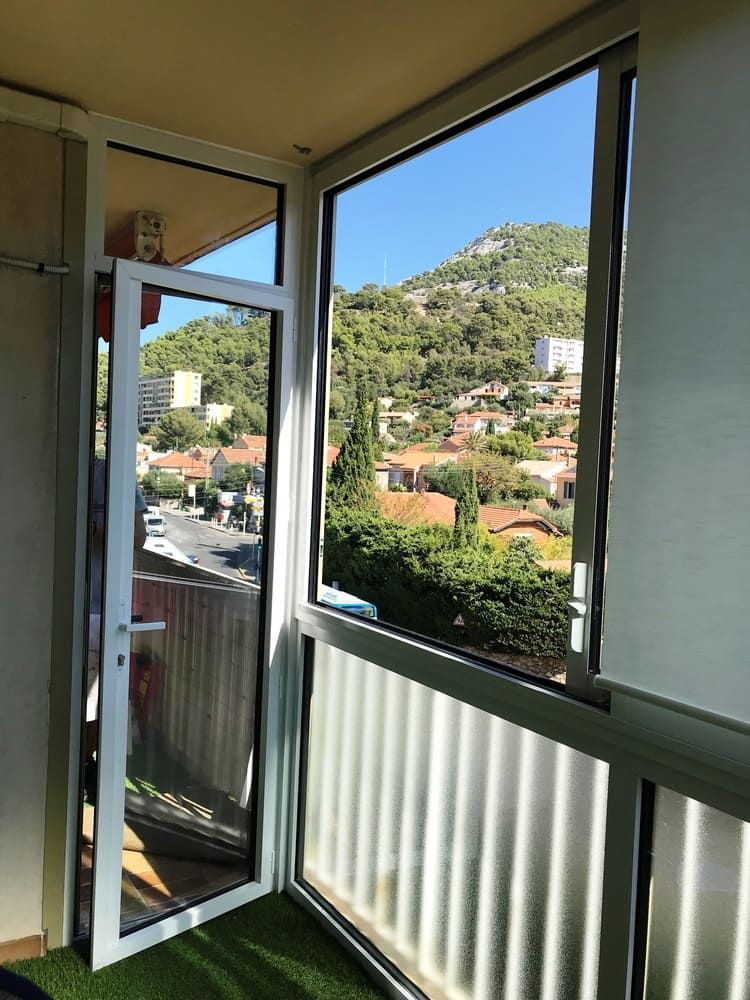 Porte blanche ouverte et fenêtre donnant sur une ville à flanc de colline et un ciel bleu.