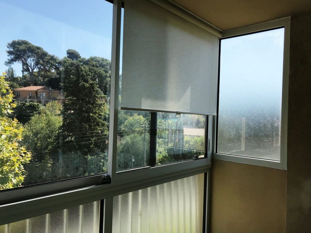 Vue d'une fenêtre ouverte avec une ombre partielle, donnant sur un paysage verdoyant avec des arbres et un bâtiment par une journée ensoleillée.