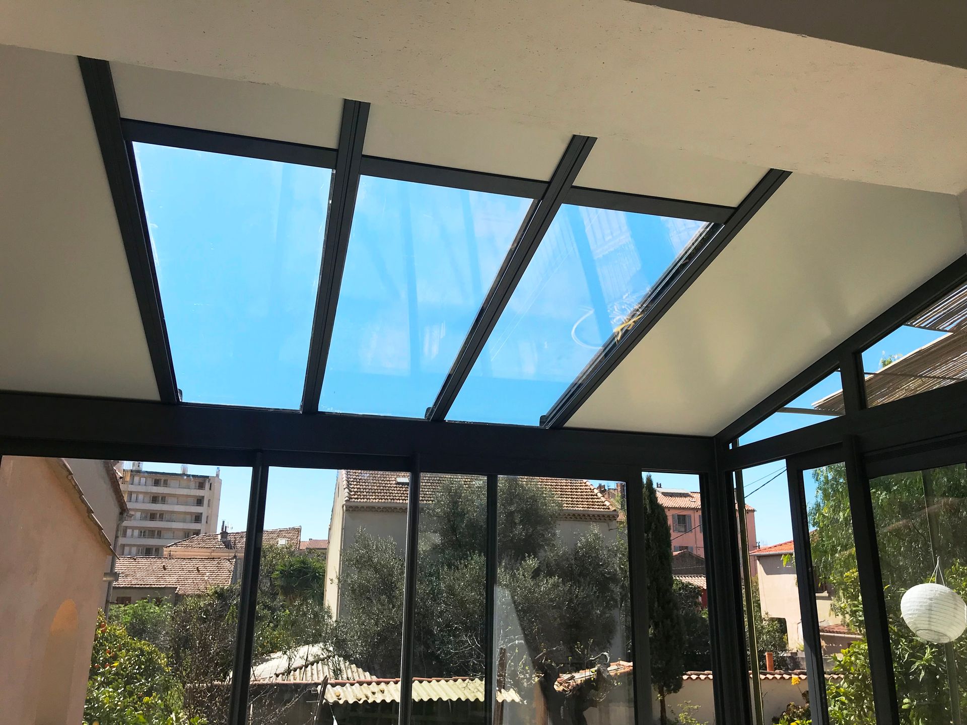 Intérieur de véranda avec toit et murs en verre, charpente noire, vue sur le ciel bleu et les bâtiments.