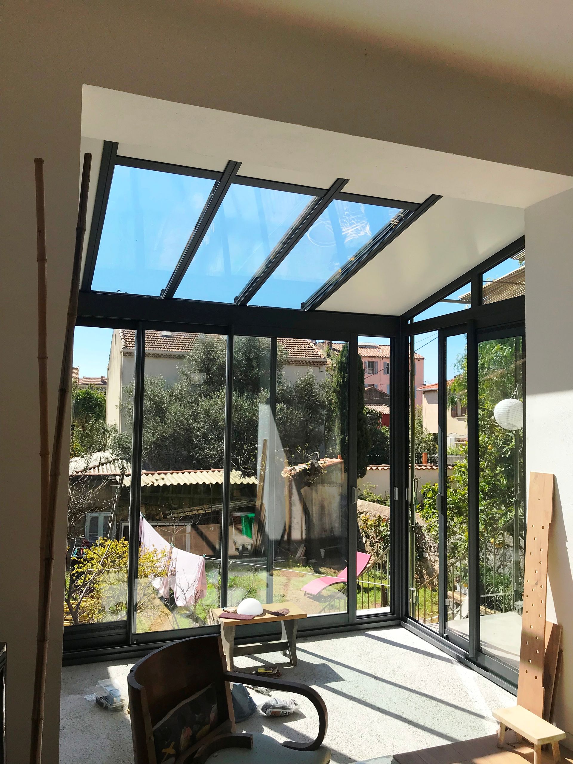 Véranda avec toit et murs en verre donnant sur un jardin. La lumière du soleil entre à flots.