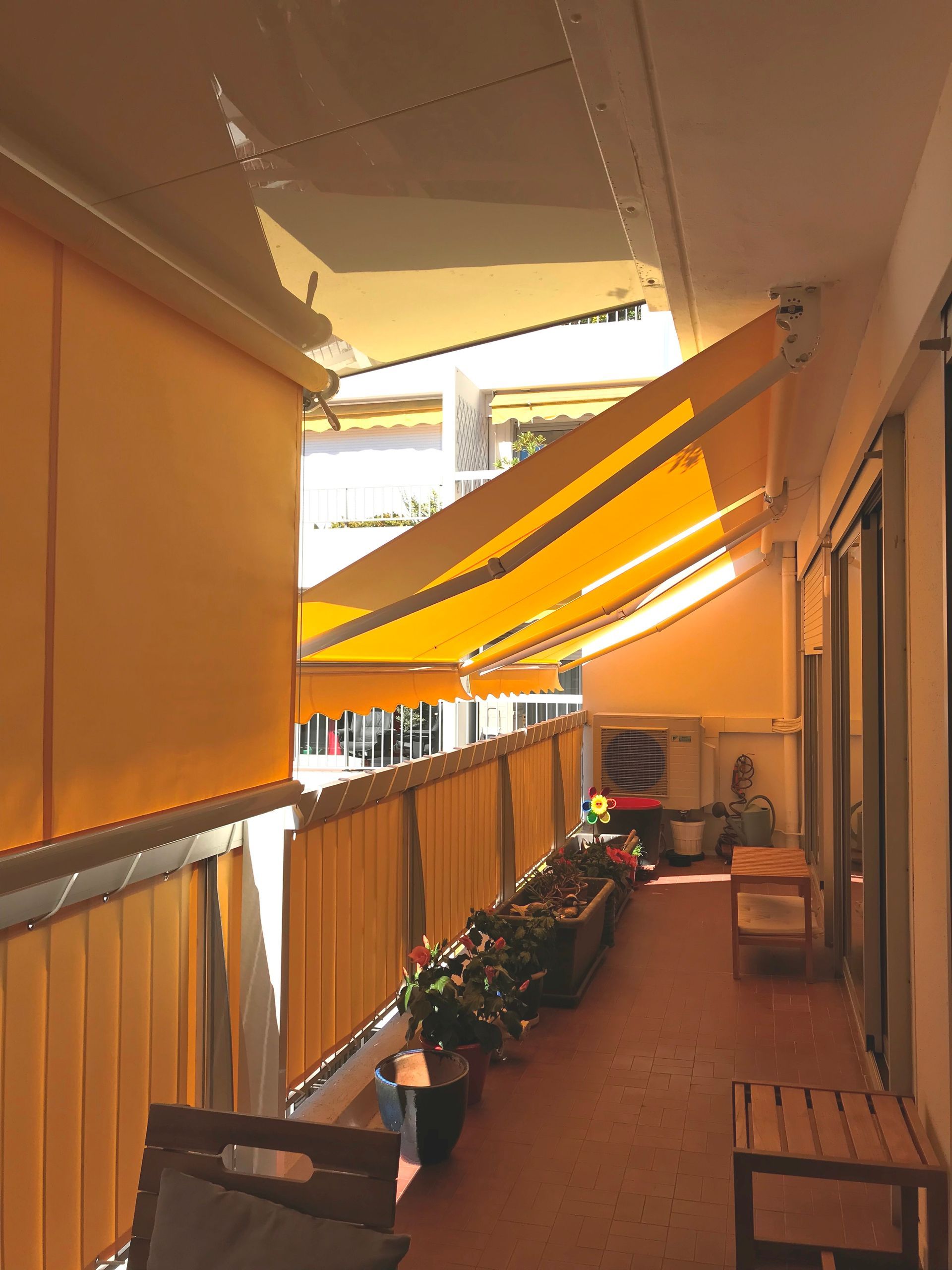 Auvents et stores jaunes sur un balcon, avec une rangée de plantes en pot et une petite table.