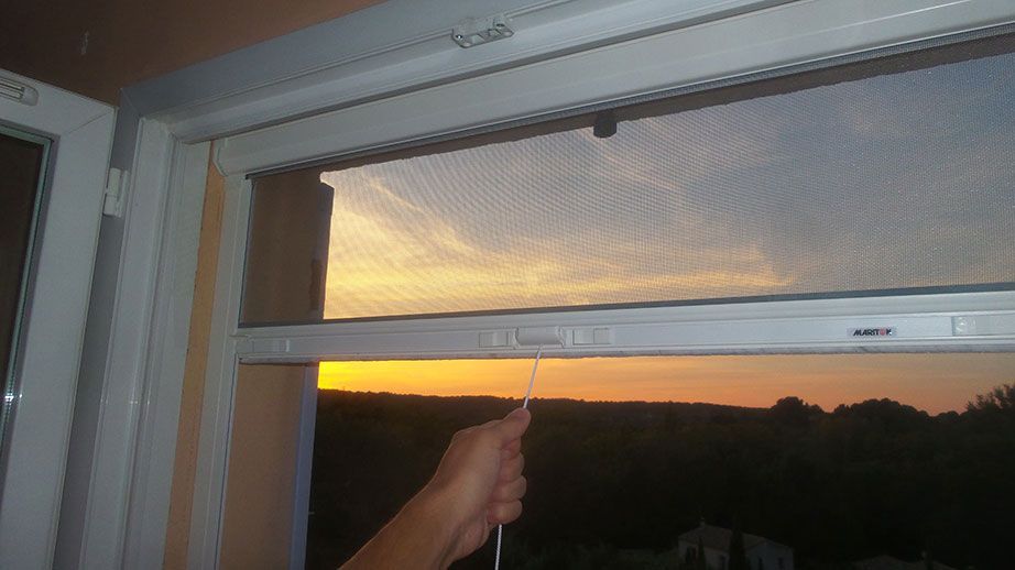 Main d'une personne tirant sur un cordon pour ajuster un écran rétractable sur une fenêtre, avec un coucher de soleil orange visible.