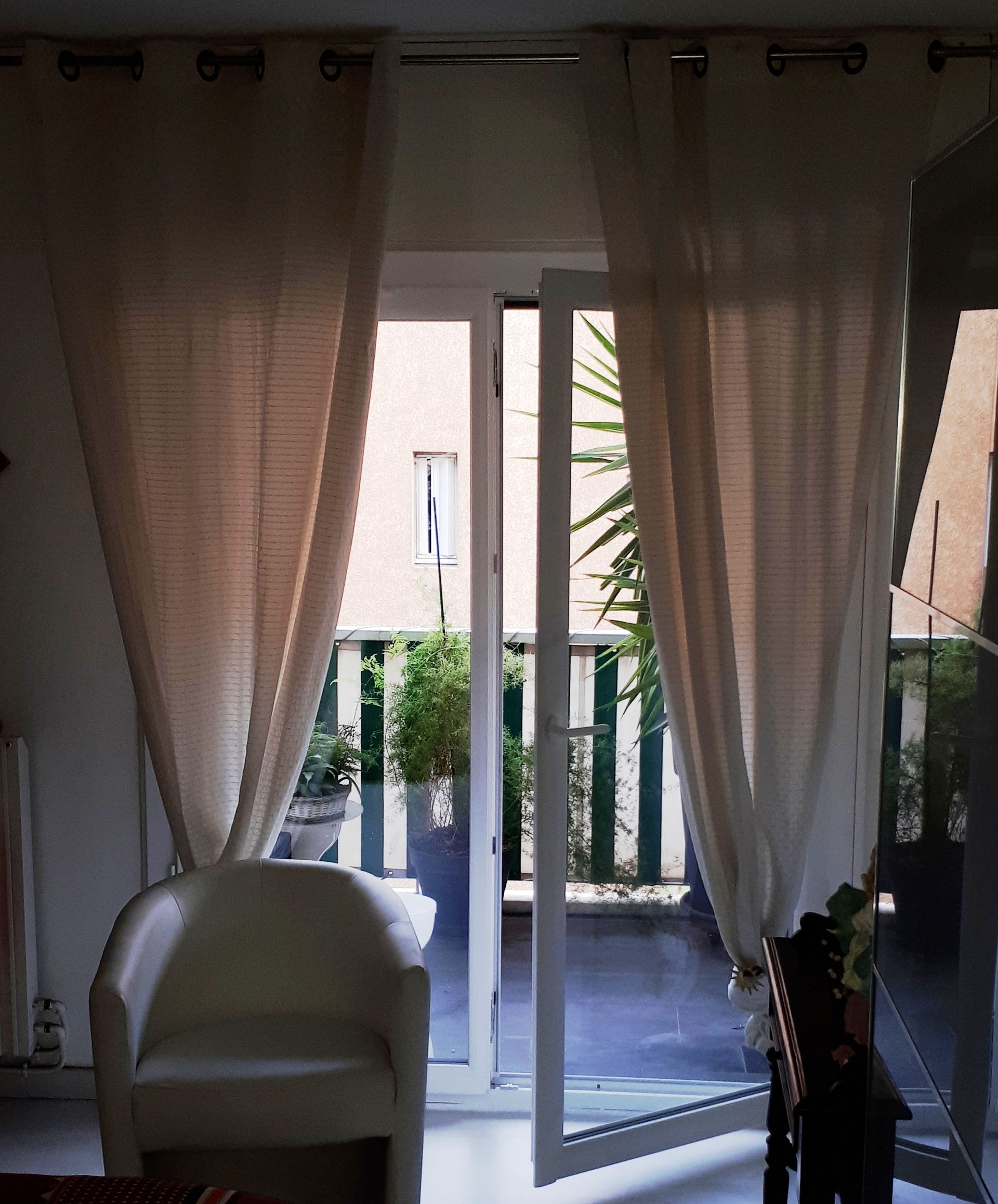 Ouvrez la porte blanche sur un balcon, avec des rideaux crème transparents encadrant la vue ; une chaise blanche se trouve à l'intérieur.