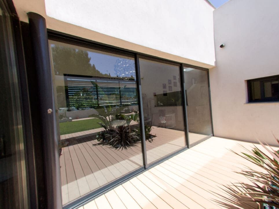 Grandes baies vitrées coulissantes sur la terrasse, avec un jardin fleuri et réfléchissant. Bâtiment blanc moderne, extérieur ensoleillé.