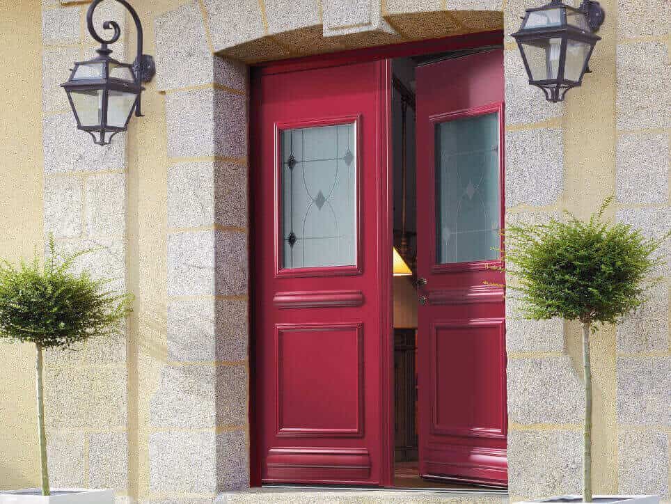 Portes doubles rouges avec panneaux de verre et fenêtres latérales, flanquées de lampes et d'arbustes taillés.