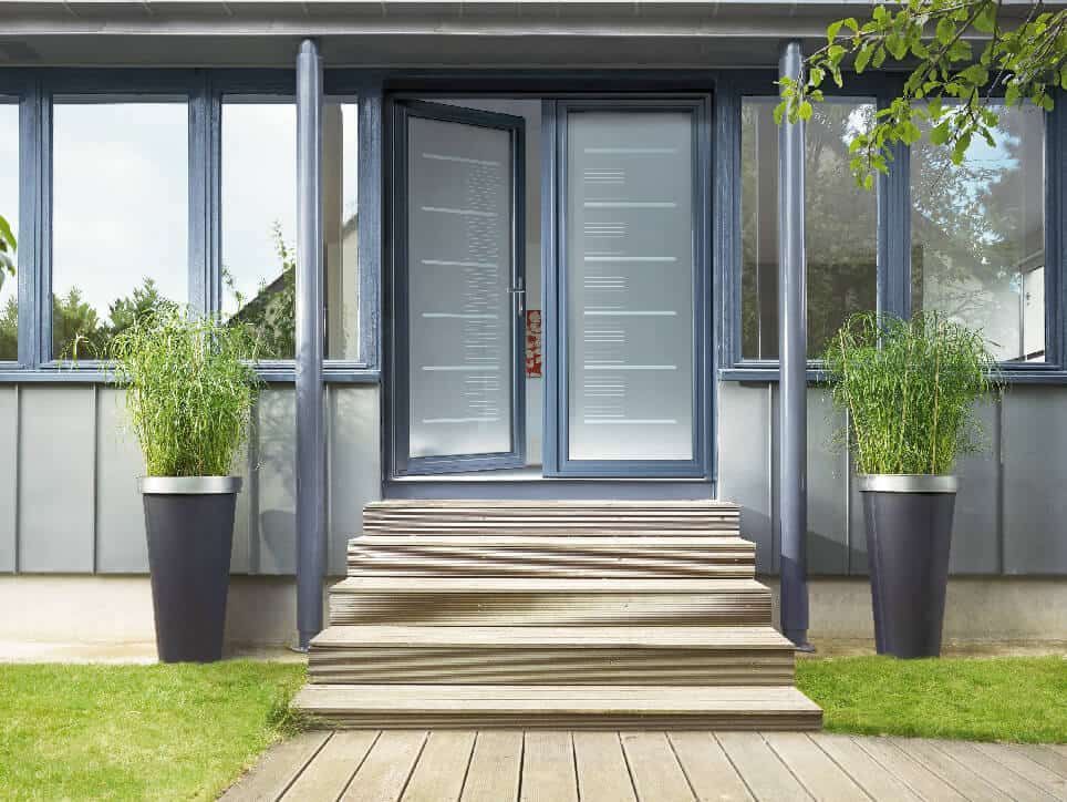 Porte d'entrée grise avec porte moustiquaire ouverte, flanquée de fenêtres et de grandes jardinières avec verdure, sur des marches en bois.
