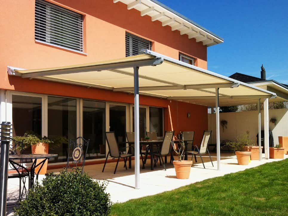 Un auvent beige recouvre une terrasse avec table et chaises. La maison est couleur pêche et possède de grandes fenêtres.
