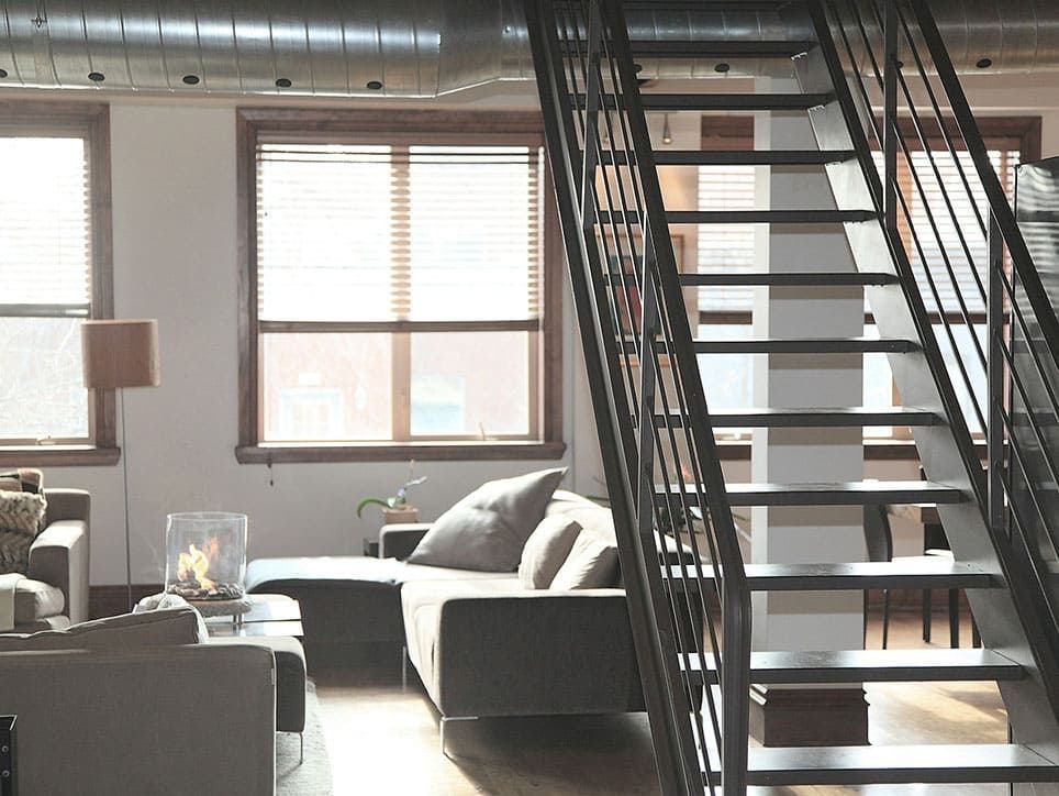 Vue intérieure d'un loft moderne avec un escalier en métal, un canapé, des fenêtres et des conduits apparents.
