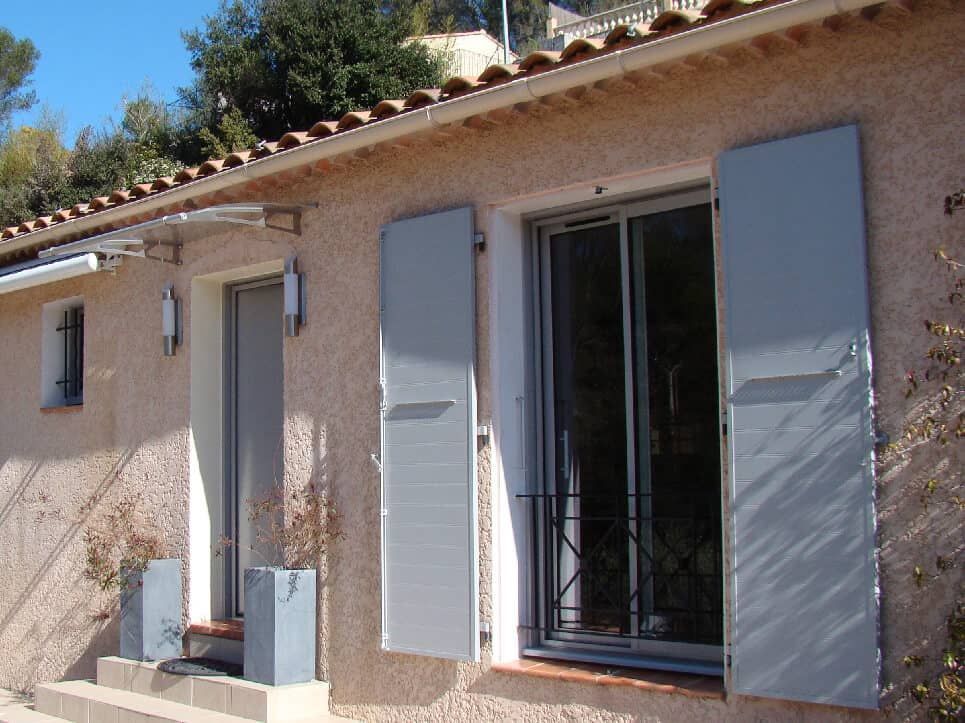 Bâtiment en stuc beige avec volets et porte gris, toit en tuiles rouges et auvent transparent.