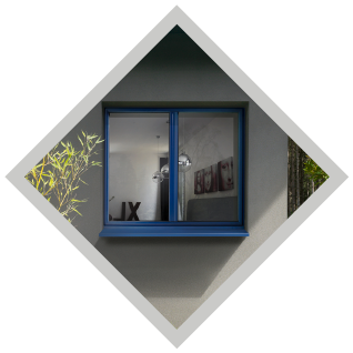 Fenêtre à cadre bleu sur un mur gris avec reflet intérieur. Bordure blanche en losange avec verdure apparente.