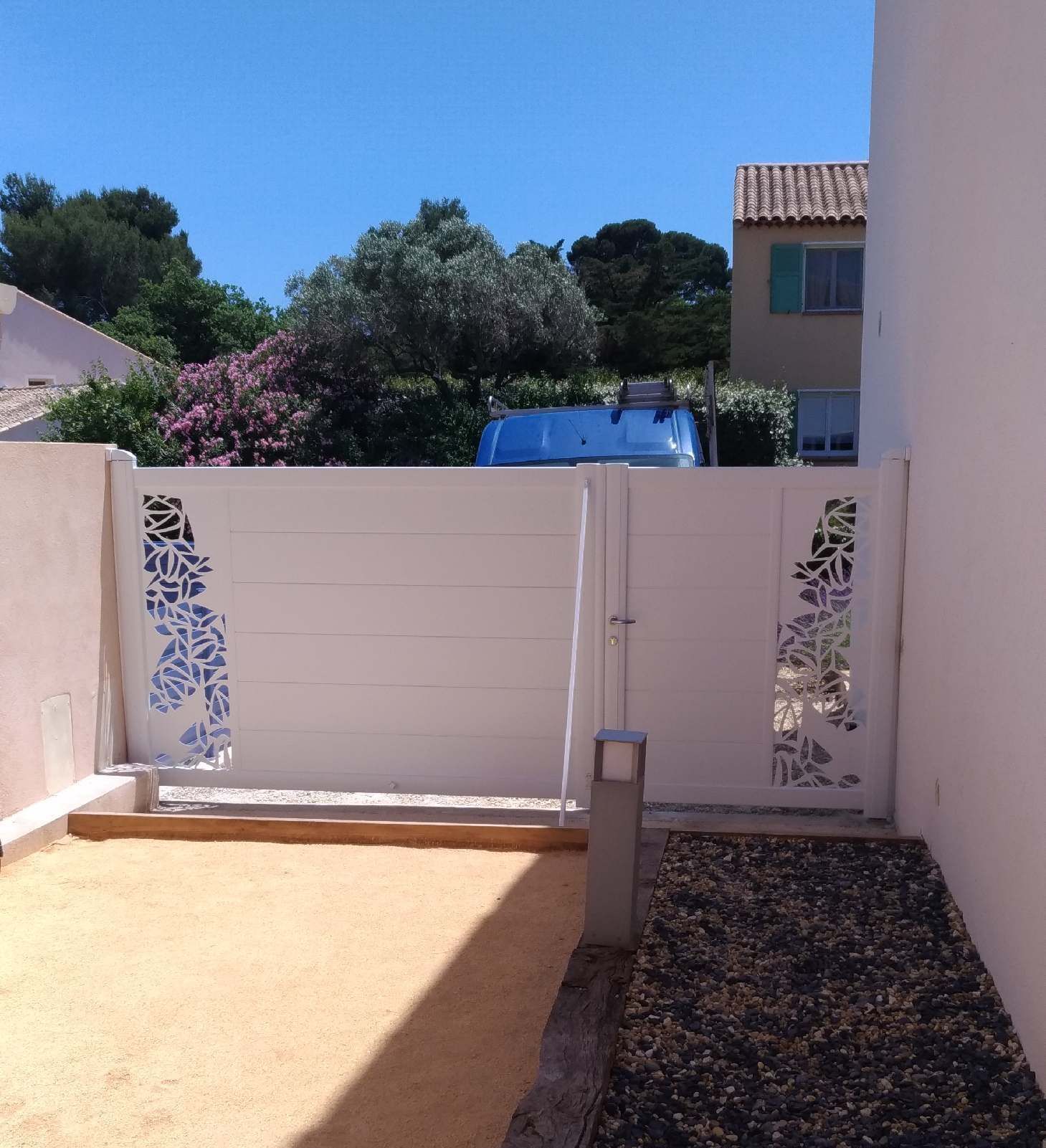 Portail blanc avec des découpes en forme de feuille, menant à une cour avec du sable brun clair et des galets foncés.