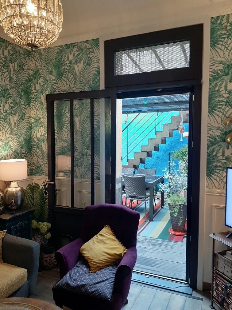 Intérieur avec papier peint à motifs, fauteuil violet et vue sur un patio bleu avec des escaliers traversant une porte.