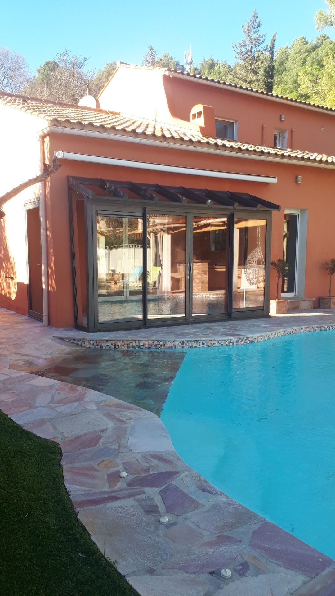 Véranda au bord de la piscine, attenante à une maison en stuc orange au toit de tuiles. Journée ensoleillée.