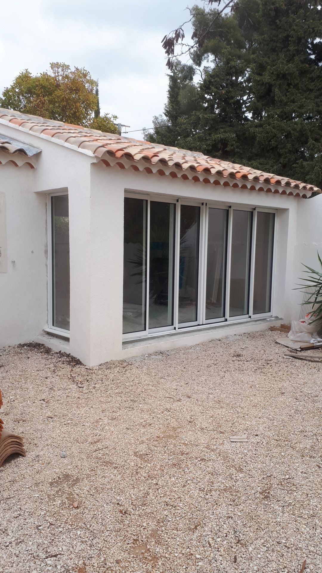 Bâtiment blanc avec toit en tuiles, portes pliantes en verre, sol en gravier et arbres environnants.