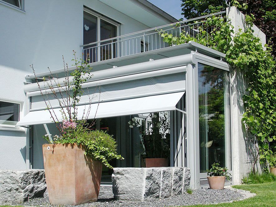 Véranda avec auvent ouvert, terrasse en pierre et balcon. Bâtiment blanc avec végétation luxuriante.