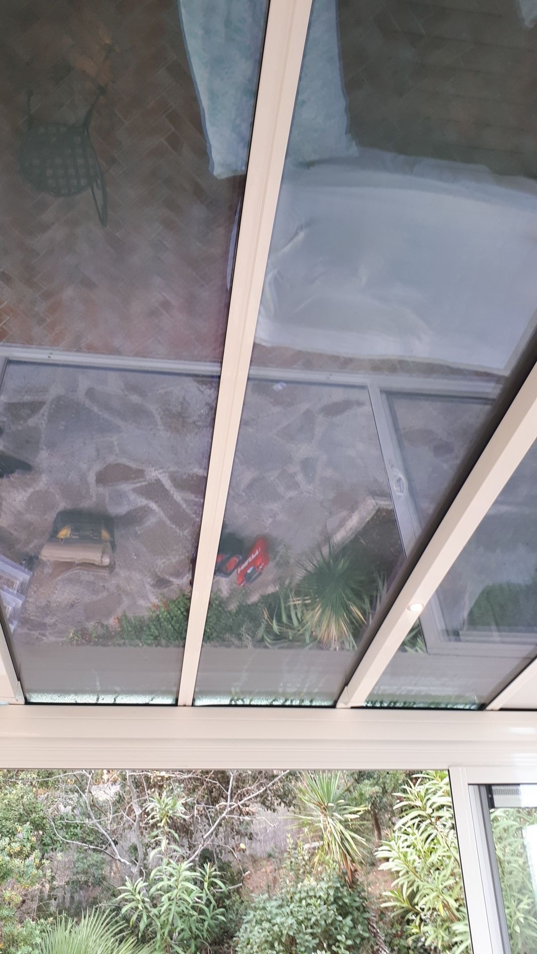 Structure à toit de verre sur un patio en pierre avec outils et herbe visibles ; cadre blanc avec verdure extérieure.
