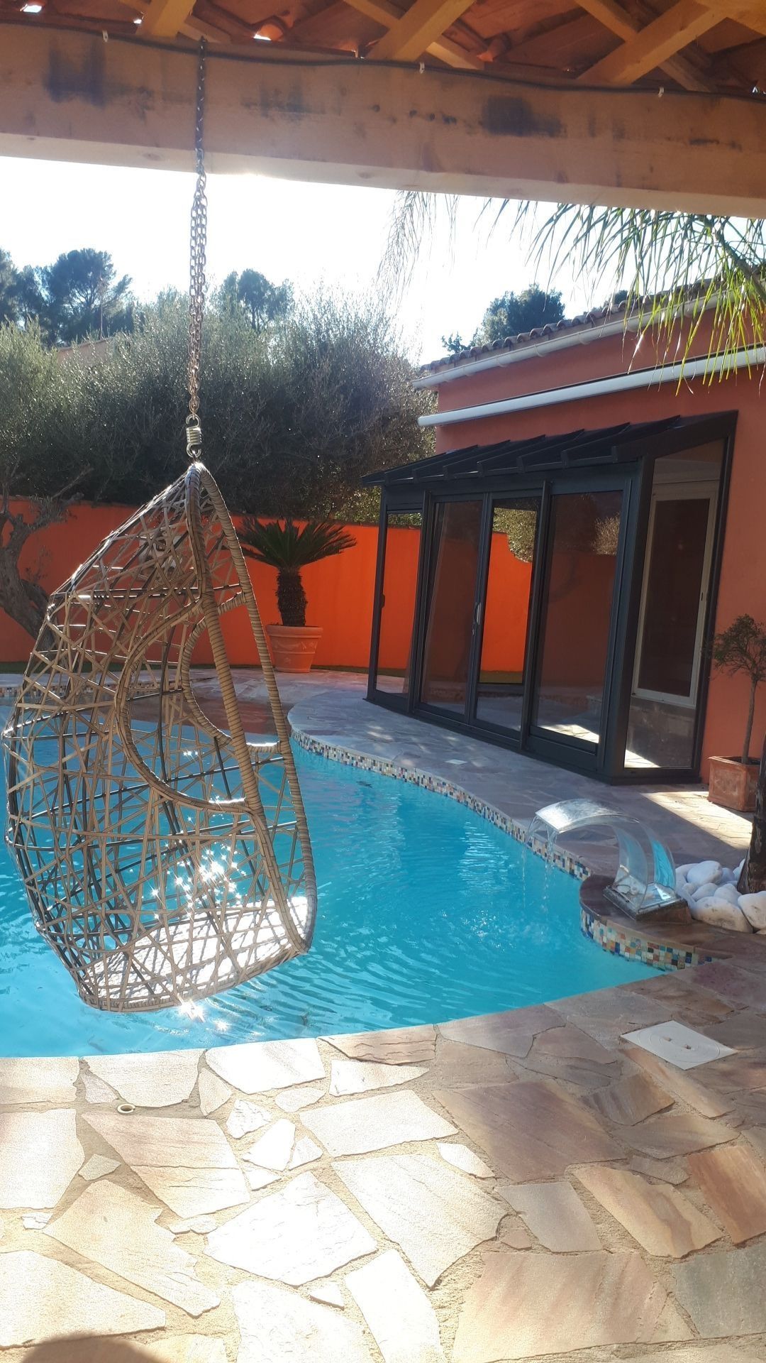 Une chaise suspendue tressée au-dessus d'une piscine ; un bâtiment rouge en arrière-plan. Cadre extérieur ensoleillé.