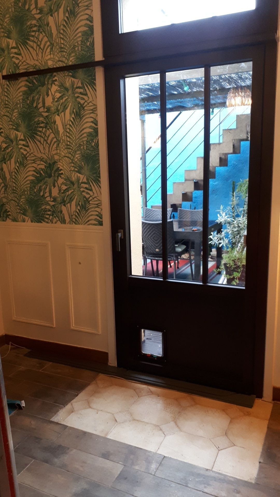 Porte marron foncé avec une chatière, des vitres et une vue sur des escaliers bleus.