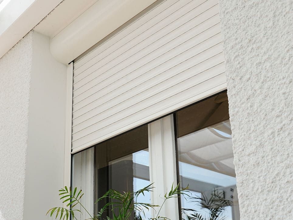 Vue extérieure d'une fenêtre avec volet roulant blanc cassé fermé. On aperçoit un mur blanc et un soupçon de feuillage.