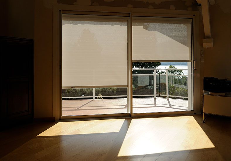 Portes coulissantes en verre avec stores beiges fermés, menant à un balcon avec soleil partiel au sol.