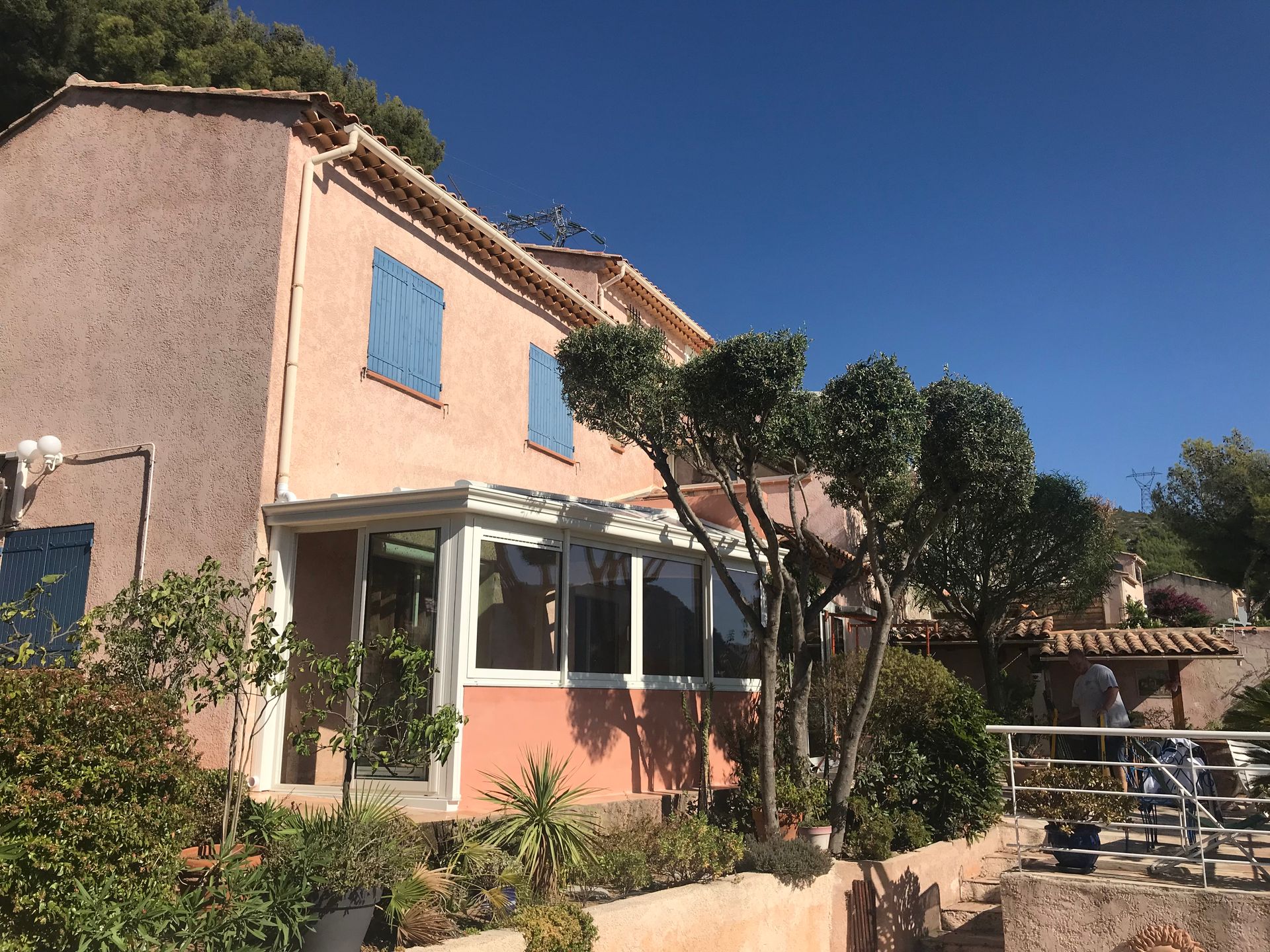 Maison en stuc rose avec des volets bleus, une véranda et un jardin contre un ciel bleu vif.