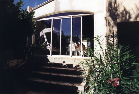 Vue extérieure d'une maison avec portes coulissantes en verre, escaliers et verdure. L'intérieur dévoile meubles et personnes.