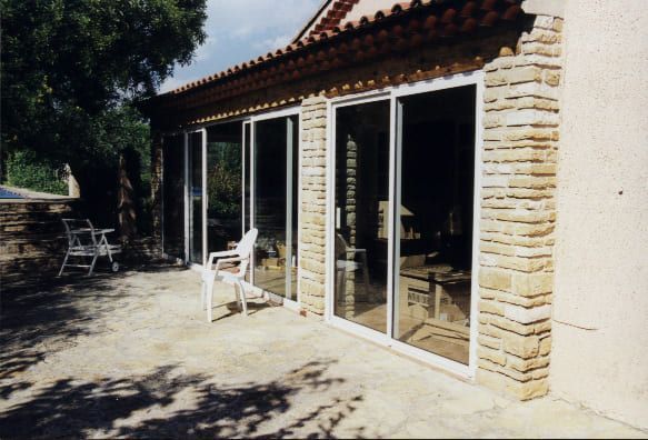 Portes coulissantes en verre sur un bâtiment aux détails en pierre et terrasse. Chaises blanches sur la terrasse.