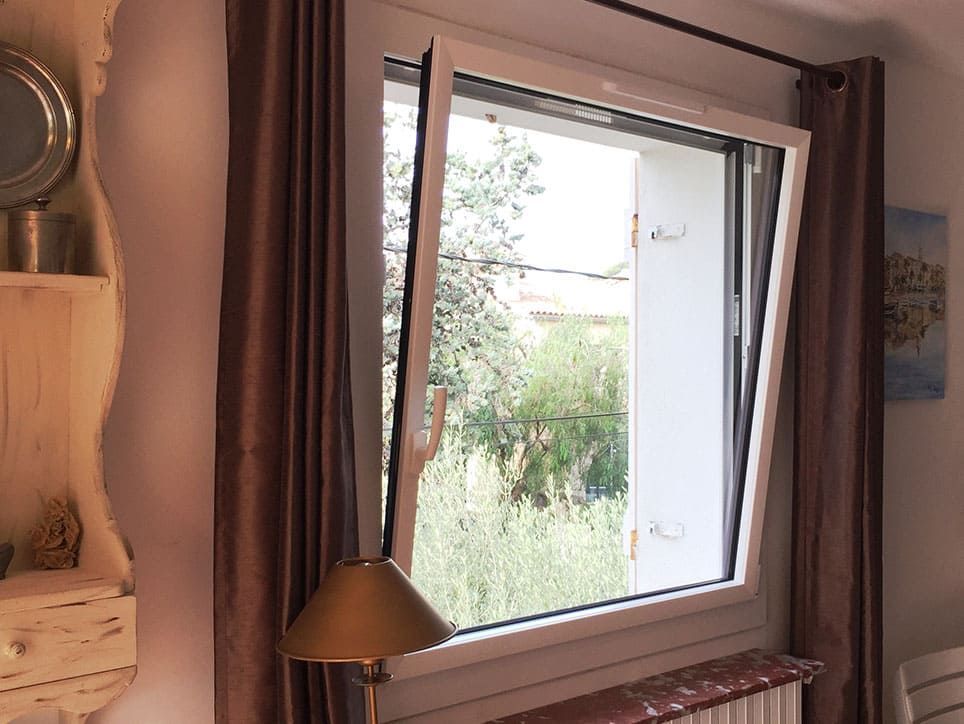 Fenêtre ouverte à cadre blanc avec vue partielle sur les arbres ; des rideaux marron encadrent la fenêtre ; une lampe se trouve en dessous.