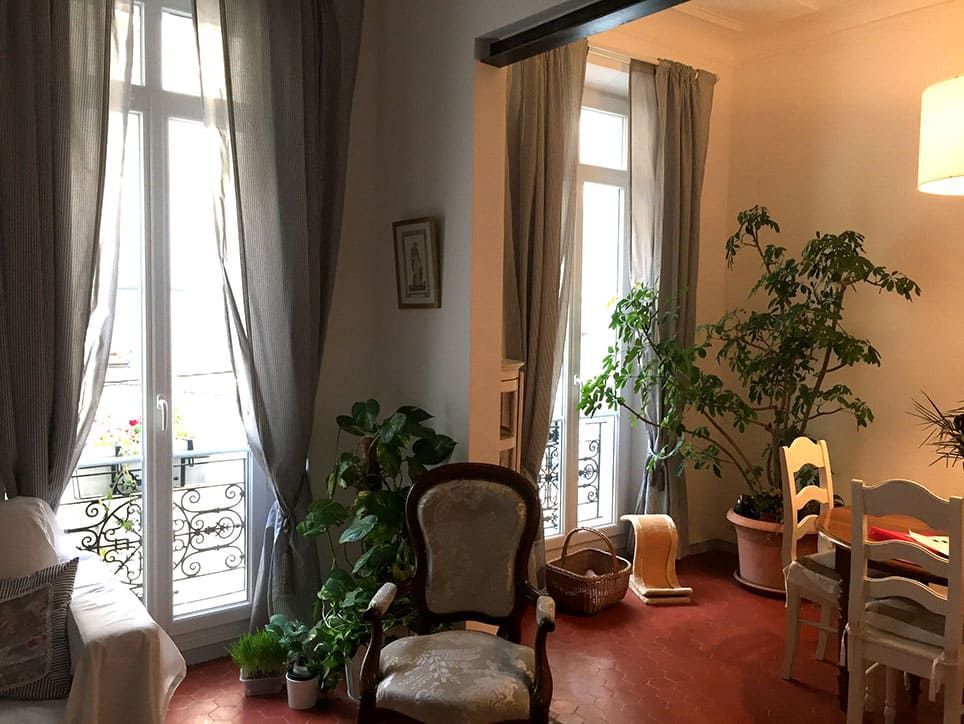 Pièce intérieure avec fenêtres ouvertes, rideaux gris transparents, plantes et meubles anciens.