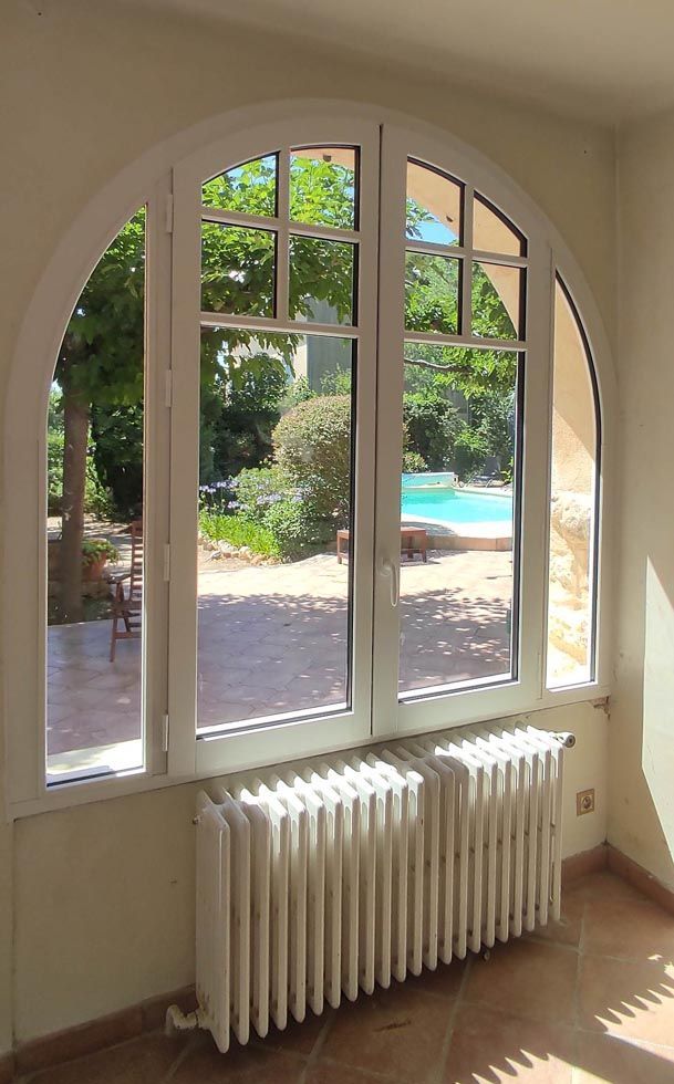 Fenêtre cintrée avec cadre blanc donnant sur un jardin et une piscine, avec un radiateur blanc en dessous.