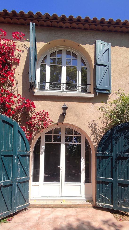 Bâtiment beige avec fenêtre et porte cintrées, volets et portails verts, fleurs rouges, ciel bleu.