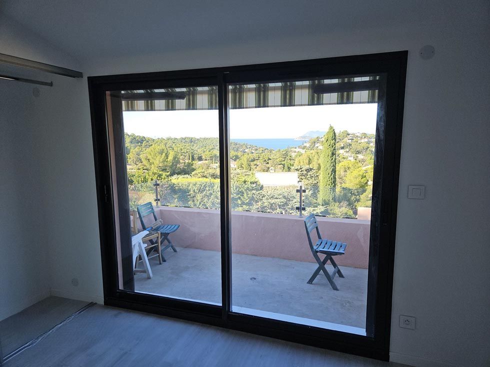 Des portes coulissantes en verre s'ouvrent sur un balcon avec des chaises, donnant sur un paysage avec des arbres et la mer.