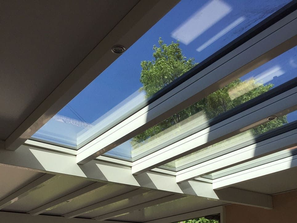 Des puits de lumière dans une terrasse couverte encadrent un ciel bleu vif et le feuillage des arbres. Des poutres blanches soutiennent des panneaux de verre.