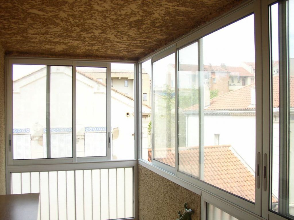Balcon entouré de fenêtres donnant sur les toits de la ville, avec des cadres blancs et un plafond texturé.