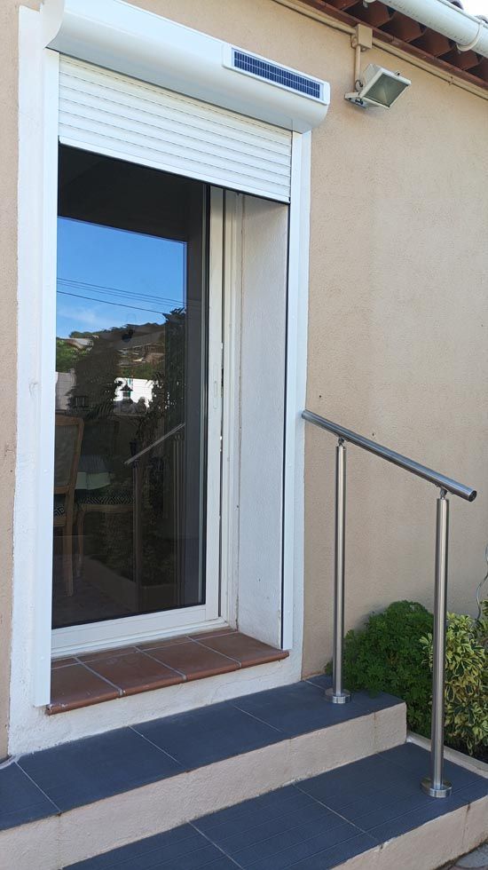 Porte extérieure avec cadre blanc, porte vitrée et volet solaire. Un escalier avec rampe mène à l'entrée.