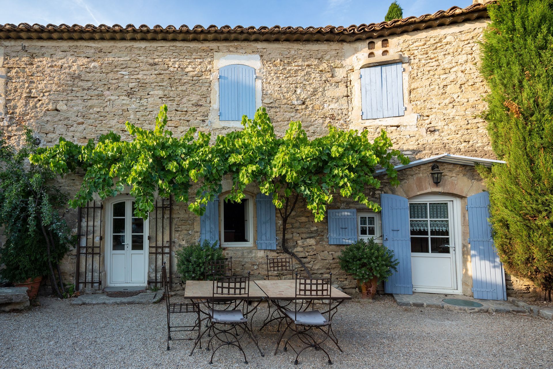 Une maison rustique en pierre avec des volets bleu clair et une treille au-dessus d'un salon de jardin