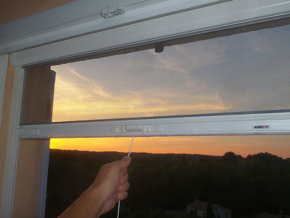 Main tirant sur un cordon pour ajuster un écran rétractable sur une fenêtre avec vue sur le coucher du soleil.