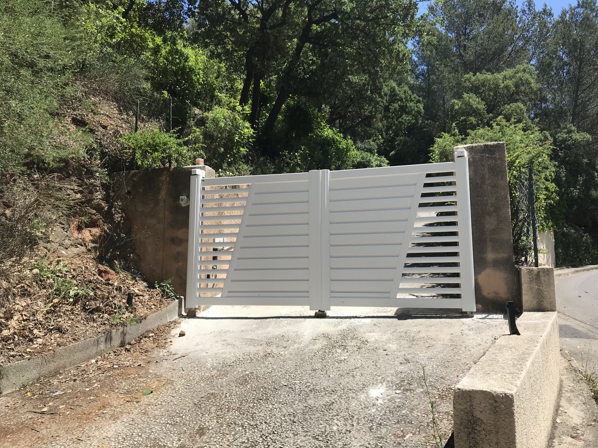 Portail d'entrée en métal blanc, à claire-voie, fermé. Murs de soutènement en béton de chaque côté, entouré de verdure.