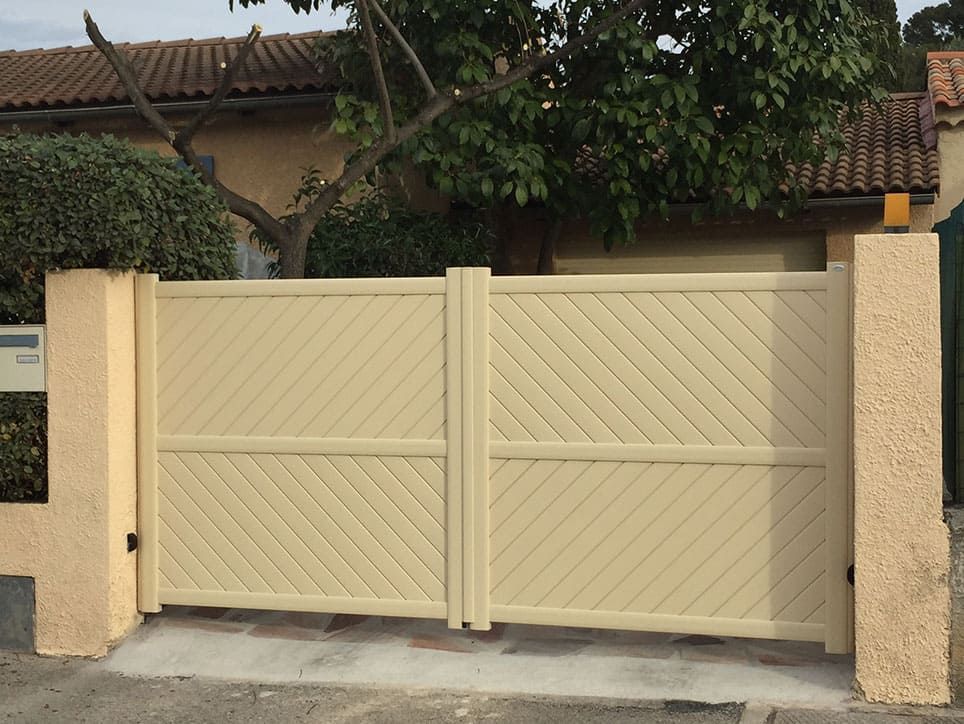 Portail double beige à motifs diagonaux devant une maison, posé sur du béton, avec des piliers de soutien de chaque côté.
